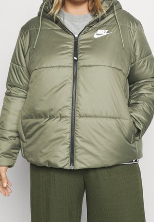 Persona con chaqueta acolchada Nike verde oliva con capucha y pantalones verdes acanalados, manos relajadas a los lados.