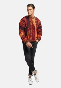 Mehrfarbiger Pullover mit einem strukturierten Muster, der orange, rot und dunkellila Farbtöne zeigt. Kombiniert mit schwarzen Jeans und weißen Sneakers.