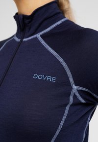 Marineblå langærmet top med høj krave og lynlås, med lys blå syning og "DOVRE" broderet på brystet. Glat stofstruktur.