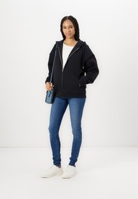 Camisola de capuz preta com fecho, um capuz grande, dois bolsos frontais, usada sobre uma camisa clara, combinada com jeans skinny azuis e ténis brancos.