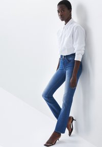 Jeans de mezclilla azul claro con corte ajustado, cintura de altura media y un ligero desvanecimiento. Combinados con una camisa blanca de botones bien ajustada y tacones negros.