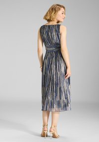 Kvinde står iført en ærmeløs, plisseret metallic midikjole med lodrette striber i blå, guld og sort, kombineret med guldhøjhælede sandaler.