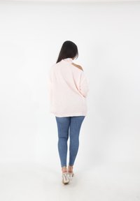 Maglione rosa chiaro oversize a collo alto con dettagli sulle spalle scoperte, abbinato a jeans blu aderenti e sandali con tacco trasparente.
