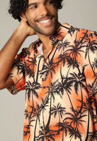 Korte mouw shirt in oranje en crème, met een zwarte palmboomprint. Gemaakt van lichte stof met een knoopsluiting en kraag.