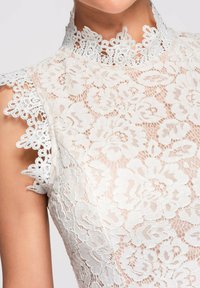 Haut sans manches en dentelle florale blanche avec un col haut festonné et des motifs brodés complexes sur tissu transparent.