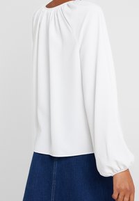 Blusa bianca a maniche lunghe con scollatura arricciata e maniche a sbuffo, realizzata in un tessuto liscio. Indossata con una gonna di jeans scuri.