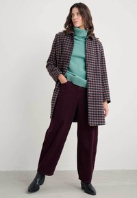 Provjeren kaput dugih rukava s gumbima, složen preko tirkiznog roll-neck pulovera. Burdeos široke pamučne hlače i crne čizme do gležnja upotpunjuju izgled.