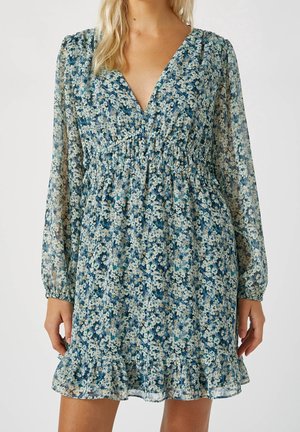 Femme portant une robe longue à manches longues bleue à motifs floraux, avec un col en V, une taille froncée et un ourlet à volants, debout devant un fond uni.