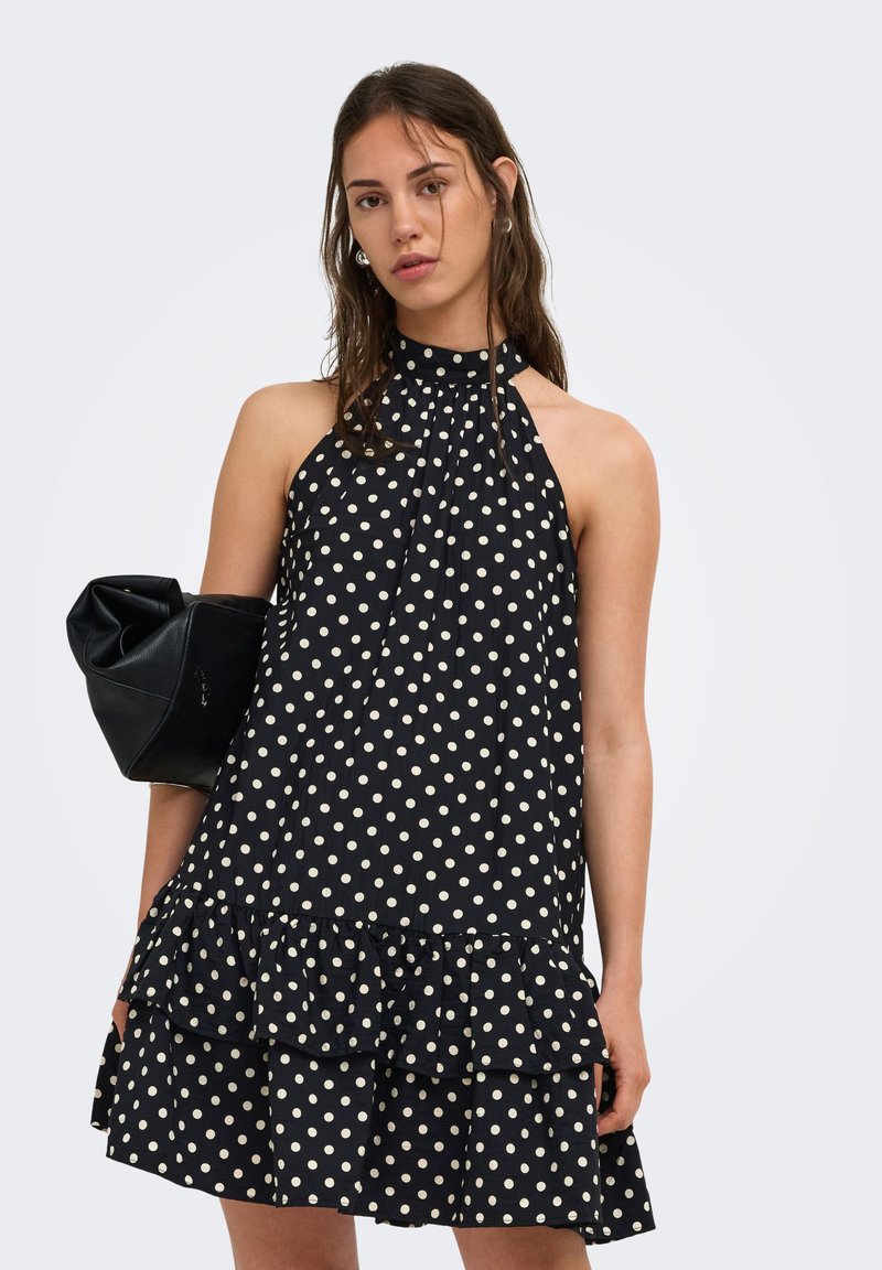 Jeune femme portant une robe noire sans manches à pois, tenant un sac à main noir sous le bras, sur un fond uni.