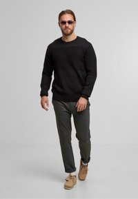 Schwarzer Strickpullover mit strukturiertem Einsatz, graue Freizeithose und hellbraune Wildlederschuhe. Die Accessoires umfassen eine leichte Sonnenbrille. Einfaches, lässiges Outfit.
