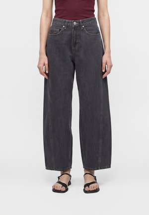 Personne portant un jean denim noir ample associé à des sandales noires à bout ouvert et un haut sans manches bordeaux, debout devant un fond blanc.