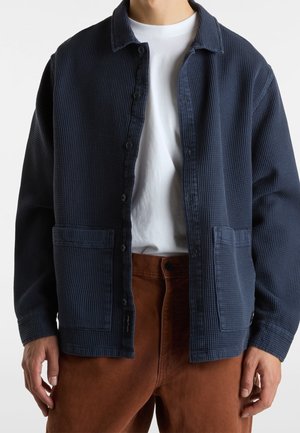 Mann trägt eine dunkelblaue Jacke mit Waffelstruktur und Knöpfen, weißes T-Shirt und braune Hose, steht mit locker an den Seiten hängen Händen.