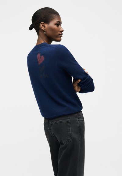 Γυναίκα με κοντά μαλλιά και χρυσούς κρίκους σκουλαρίκια, φορώντας μπλε πουλόβερ με κόκκινη καρδιά και κείμενο στην πλάτη, συνδυασμένο με μαύρα τζιν.