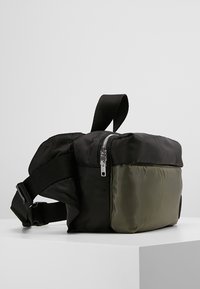 Borsa in nylon nera e verde oliva con forma rettangolare, tracolla regolabile, zip superiore e texture liscia. Presenta una parte laterale nera prominente.