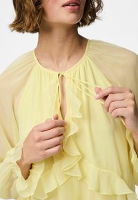 Blusa amarilla ligera con escote con volantes y cierre frontal con lazo. Cuenta con mangas largas y fluidas y una textura transparente.