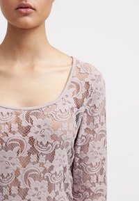 Top de encaje con escote en forma de barco, con un patrón floral en suave lavanda, con textura transparente y mangas largas.