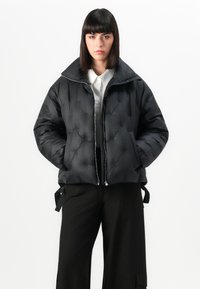 Giacca a vento nera con design trapuntato, colletto alto, chiusura con zip e tasche laterali. Indossata sopra una camicia bianca e pantaloni neri.