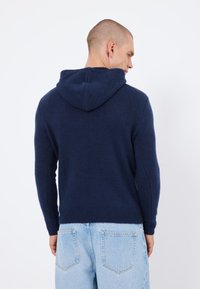 Mörkblå huvtröja med texturerad stickning, åtsittande ärmslut och ribbad nederkant. Bärs med ljusblå jeans, som visar bakvy.