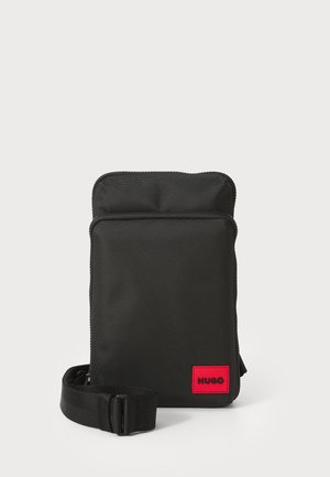 Sac à bandoulière noir en nylon avec une texture lisse, de forme rectangulaire, fermeture zippée et un patch logo rouge à l'avant. Sangle ajustable incluse.