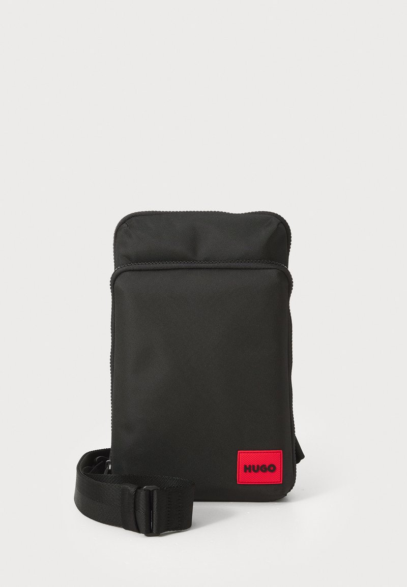 Schwarze Nylon-Crossbody-Tasche mit glatter Textur, rechteckiger Form, Reißverschluss und einem roten Logo-Patch auf der Vorderseite. Verstellbarer Trageriemen enthalten.