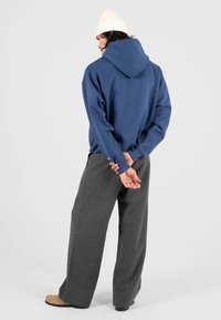 Sudadera azul con un gran bolsillo trasero, combinada con pantalones de pierna ancha grises y un gorro de punto crema. Textura suave, corte relajado.