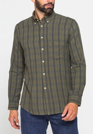 Uomo che indossa una camicia a quadri verde e blu con tasca sul petto e jeans blu su sfondo neutro.