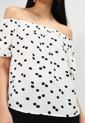 Woman wearing a white off-shoulder blouse with scattered black polka dots and short sleeves, paired with black bottoms.