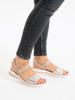 Pies calzados con sandalias beige de punta abierta con patrones geométricos y esmalte de uñas rojo, combinados con jeans ajustados gris oscuro sobre un fondo blanco.