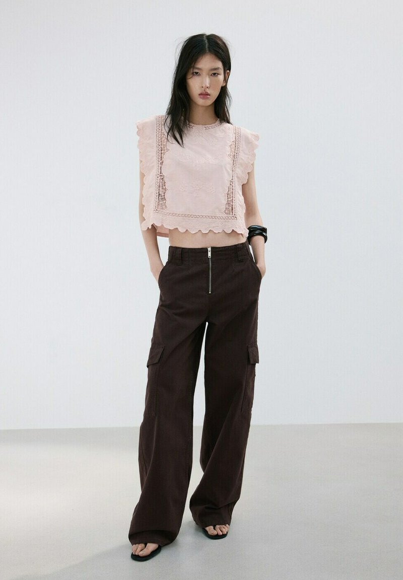 Jeune femme portant un haut sans manches en dentelle rose pâle et un pantalon cargo noir à jambes larges, debout les mains dans les poches.