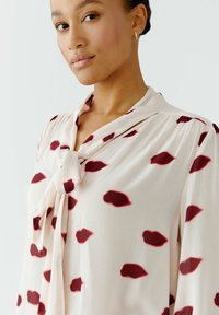 Cremfarbige Bluse mit burgunderroten Blattmustern, die über ein gebundenes Kragen-Detail verfügt und aus einem weichen, leichten Stoff besteht. Strukturierte Schultern, fließende Ärmel.
