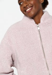 Šviesiai rožinė fliso striukė su aukštu apykakle ir sidabriniu užtrauktuku. Audinys atrodo minkštas ir tekstūruotas, tinkamas kasdieniam nešiojimui.