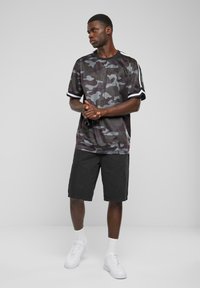 Camisa de manga corta con patrón de camuflaje negro y acentos blancos, combinada con pantalones cortos negros y zapatillas blancas, sobre un fondo neutro.