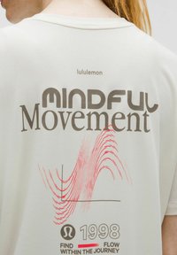 Rückseite eines cremefarbenen Lululemon-Shirts mit dem Schriftzug "Mindful Movement", einem abstrakten roten Wellenmuster, "1998" und dem Text "Find Flow Within the Journey".