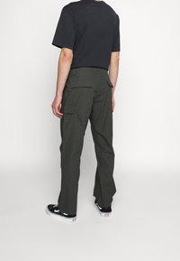 Person som bär mörkgrå cargobyxor med fickor med lock och en svart t-shirt, stående mot en enkel ljus bakgrund.