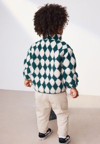 Giacca sfocata con motivo a losanghe verde scuro e bianco, abbinata a pantaloni beige e sneakers nere. Tessuto morbido e vestibilità rilassata.