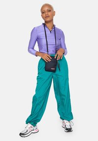 Maglietta viola a maniche lunghe con zip; pantaloni azzurro chiaro a vestibilità morbida; borsa a tracolla nera con logo; sneakers bianche e rosa. Tessuti morbidi, vestibilità rilassata.