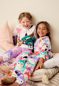Två barn sitter på en säng och har på sig färgglada pyjamaser. En har en vit överdel med djurtryck och rosa byxor, den andra har en mönstrad överdel och lila byxor.