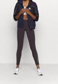 Mujer ajustando una sudadera con cremallera de Adidas en color azul marino, vistiendo un sujetador deportivo blanco, leggings morados oscuros con franjas reflectantes y zapatillas blancas de Adidas.