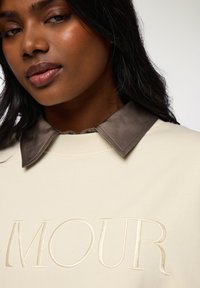 Felpa beige con design ricamato "AMOUR", caratterizzata da un colletto in satin marrone a contrasto. Tessuto liscio e vestibilità comoda.