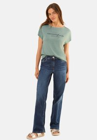 T-shirt vert clair à manches courtes avec une finition texturée et un texte imprimé, associé à un jean bleu à coupe droite et des sandales beiges.