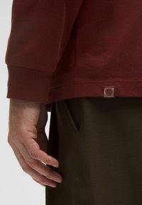 Cămașă burgundy cu mânecă lungă, cu mansete ribbed, cusături vizibile și un eticheta de accent la margine. Purtați cu pantaloni verzi măslini.