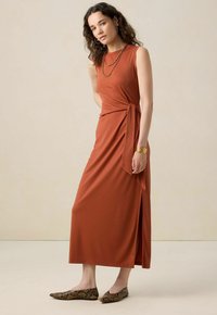 Robe sans manches de couleur rouille avec un design portefeuille et une fente sur le côté. Associée à des ballerines à motifs et des colliers superposés. Texture du tissu lisse.