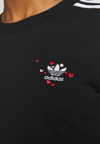 T-shirt nera in cotone con logo Adidas ricamato bianco e piccoli cuori rossi e rosa nella zona sinistra del petto.