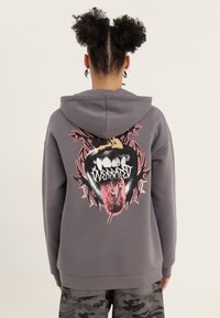 Sudadera con capucha gris con un diseño gráfico en la espalda de una boca con una lengua y púas, con acentos en rosa y negro. Tejido suave, ajuste holgado.