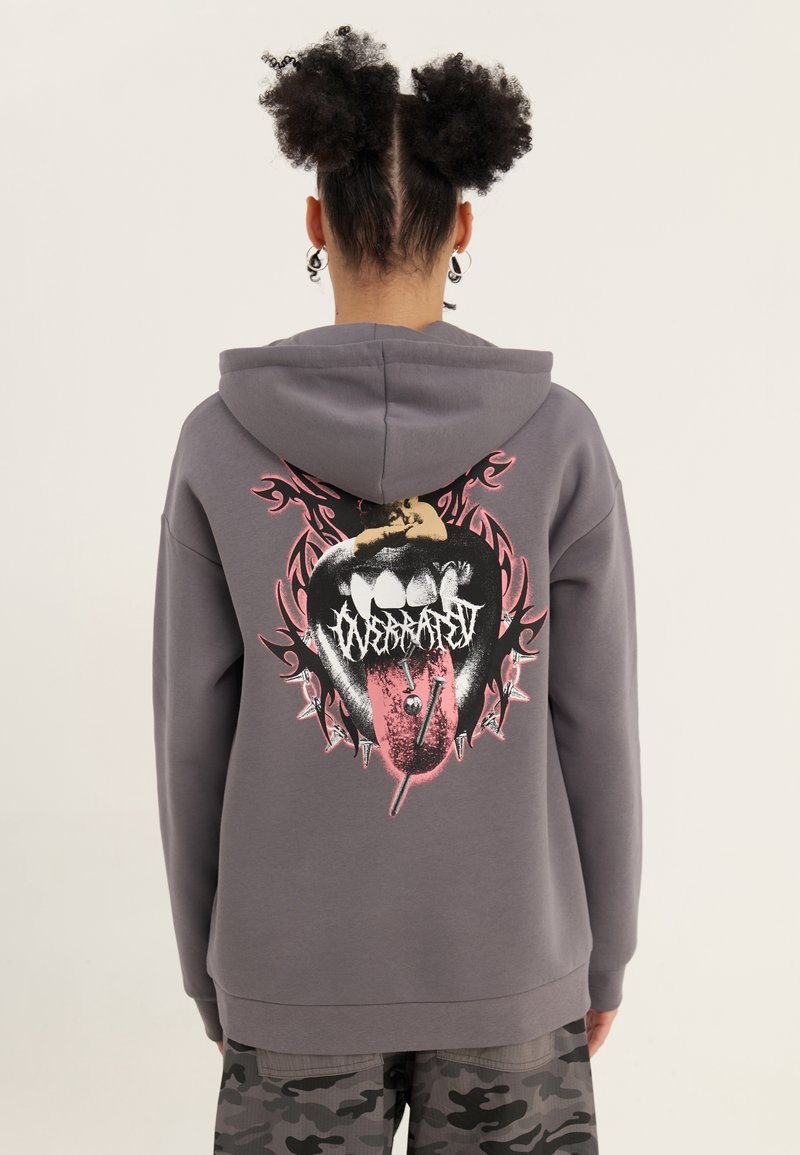Sudadera con capucha gris con un diseño gráfico en la espalda de una boca con una lengua y púas, con acentos en rosa y negro. Tejido suave, ajuste holgado.