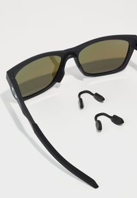 Schwarze Sonnenbrille mit grün getönten Gläsern, ausgestattet mit einem stabilen rechteckigen Rahmen und strukturierten Bügeln. Enthält zwei austauschbare Nasenpolster.