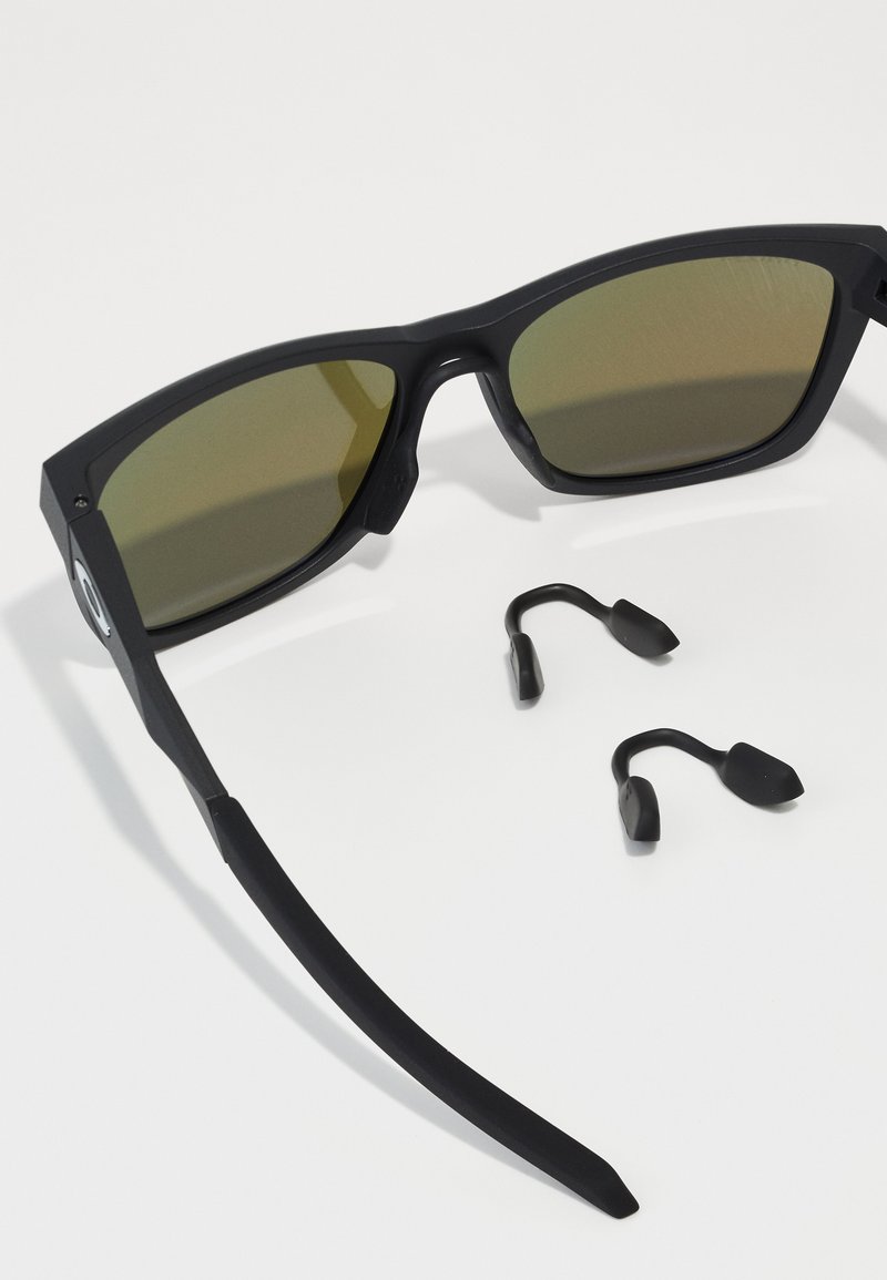 Occhiali da sole neri con lenti di colore verde, dotati di una robusta montatura rettangolare e templi con texture. Include due poggiaponte intercambiabili.