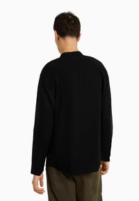 Camicia nera a maniche lunghe con colletto, caratterizzata da una texture semplice e polsini con bottoni, abbinata a pantaloni verde oliva visibili in vita.