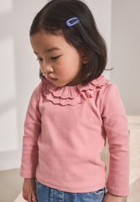 Roze top met lange mouwen en een gerimpelde halslijn met kanten rand, gecombineerd met blauwe spijkerbroek. Heeft een paarse haarclip in donker haar.