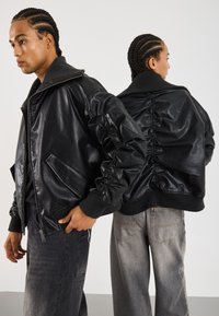 To personer med flettehår som har på seg svarte skinnbomberjakker og grå jeans, vendt mot hverandre mot en ensfarget bakgrunn.